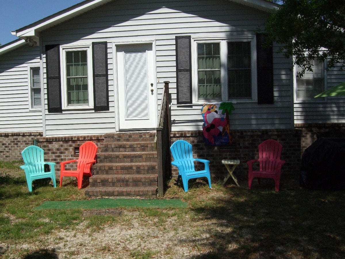 The exterior showcases a charming entrance with a set of steps leading up to the front door. Colorful Adirondack chairs in pink and blue are arranged on a grassy area, accompanied by a small table, creating a welcoming outdoor seating space.
