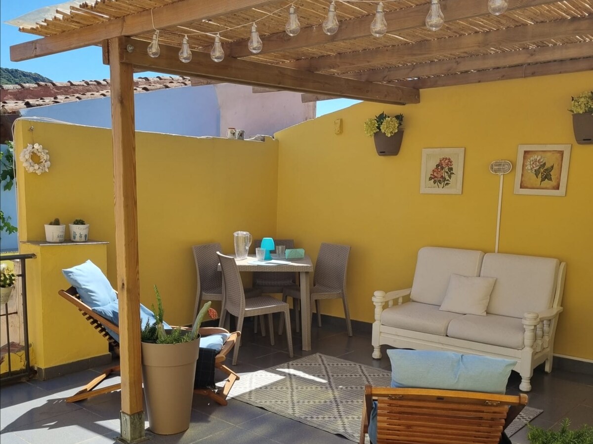 A bright terrace features a cozy seating area with a white sofa and several wicker chairs. A dining table is set for four under a wooden pergola, with soft lighting and decorative plants adorning the walls, creating an inviting space for relaxation and dining.