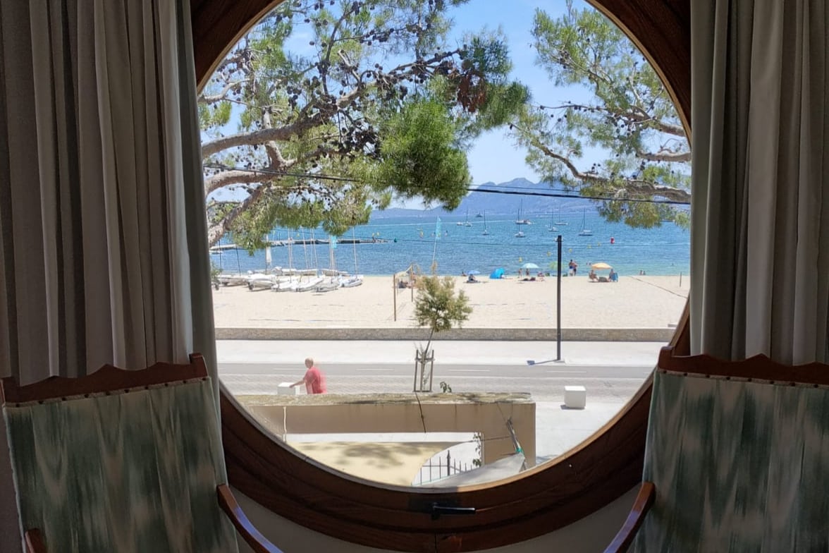A round window frames a scenic view of the beach at Puerto de Pollensa. Gentle waves lap at the shore, with boats visible on the water. Lush trees provide shade, while a pedestrian strolls along the vibrant waterfront promenade.