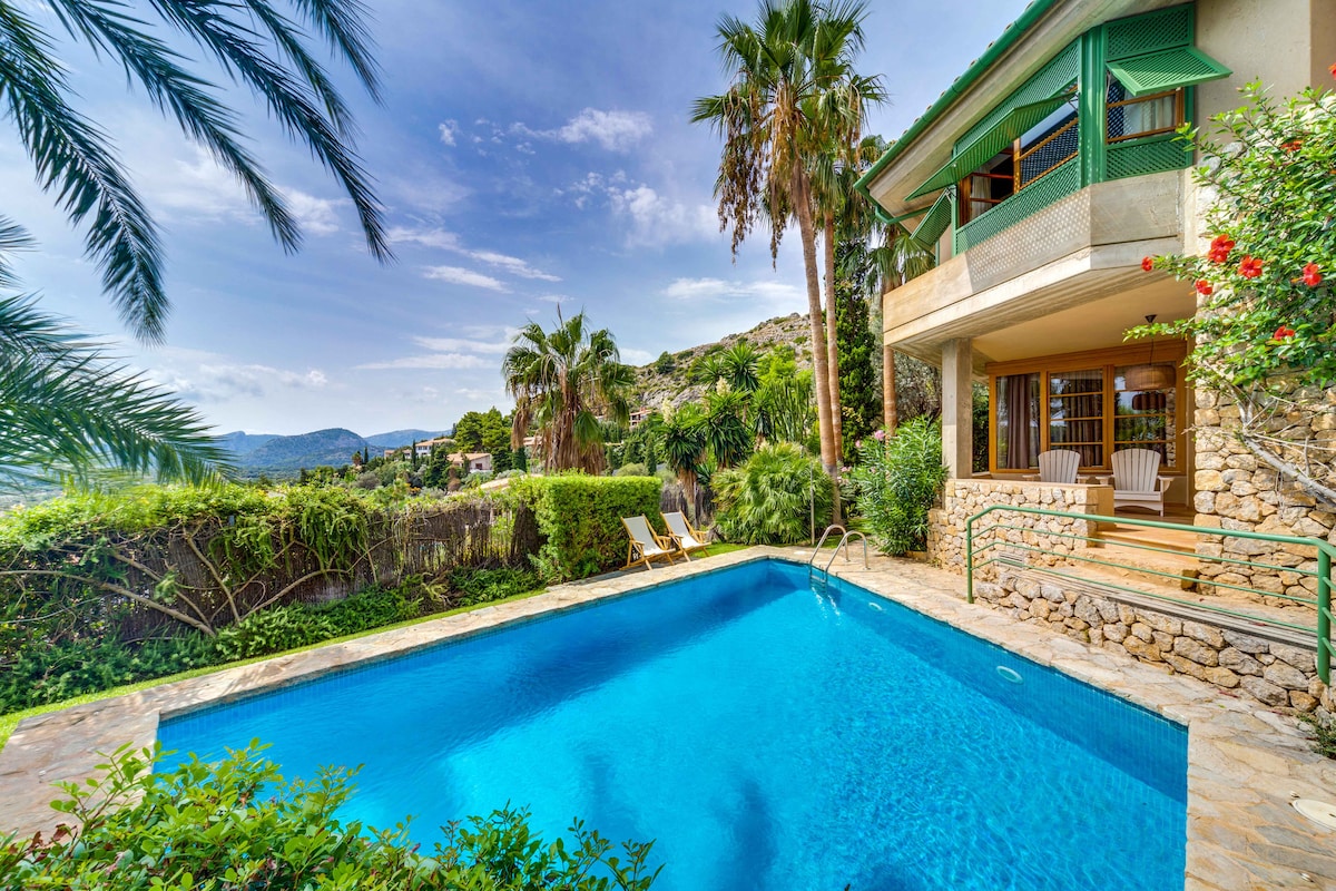 The outdoor area features a vibrant swimming pool surrounded by lush greenery and palm trees. Sun loungers are positioned beside the pool, inviting relaxation. The villa stands nearby with large windows that open to the picturesque landscape of hills and trees.