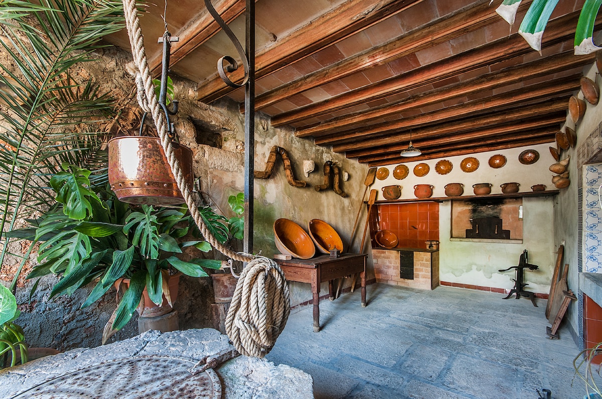 A rustic kitchen space features traditional copper cookware hanging on the walls, with a large stone table positioned in the center. Lush greenery is present, adding to the charm of the room, while terracotta tiles line the cooking area against a simple backdrop.