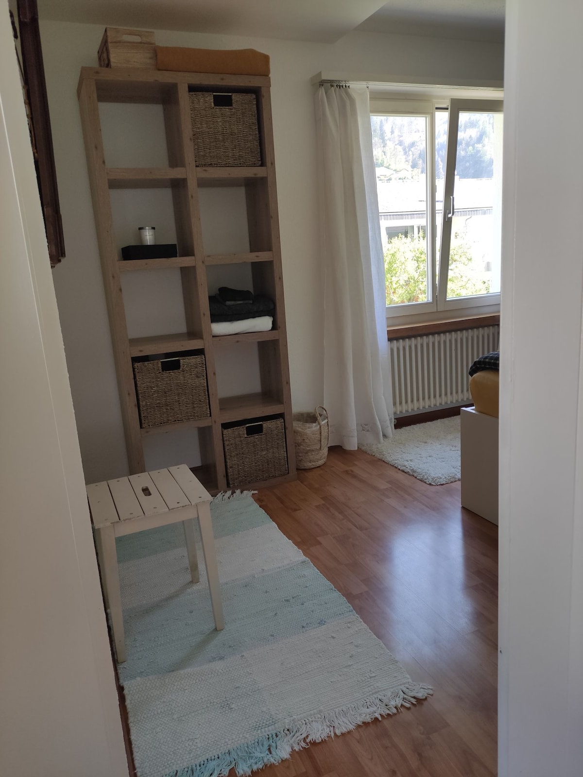 A well-lit room features a wooden shelving unit with woven storage baskets and an assortment of items. A small table stands on a light rug, while a window allows natural light to brighten the space. Light wood flooring complements the neutral decor.