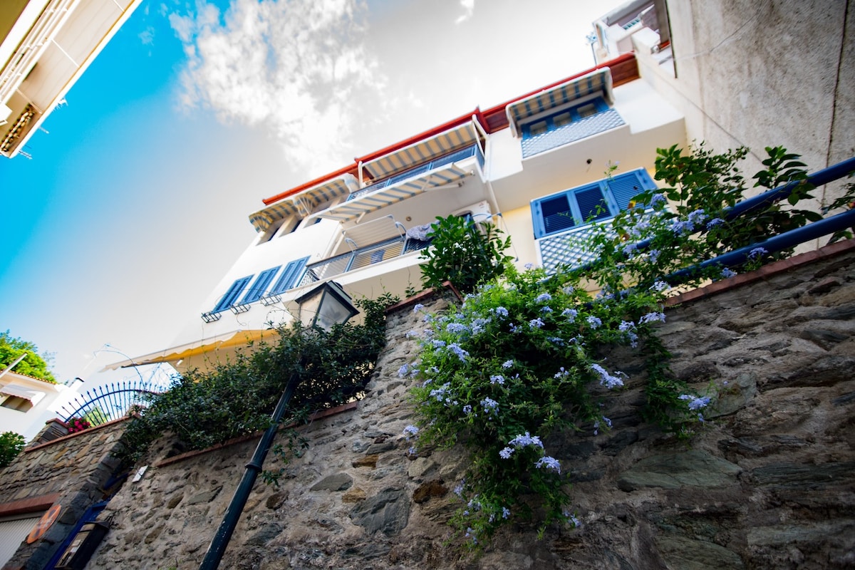 A traditional-style building is observed from below, featuring a stone wall adorned with lush greenery and flowering plants. Blue shutters frame the windows, and vibrant flowers add a touch of color. A clear blue sky with soft clouds is visible above.