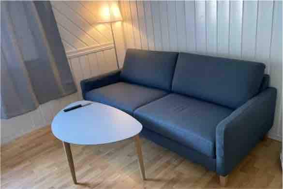 A comfortable gray sofa is positioned against a light-colored wall, accompanied by a small round coffee table. Soft lighting is provided by a nearby lamp, creating a relaxed atmosphere. A pair of sheer curtains allows natural light to gently filter into the space.