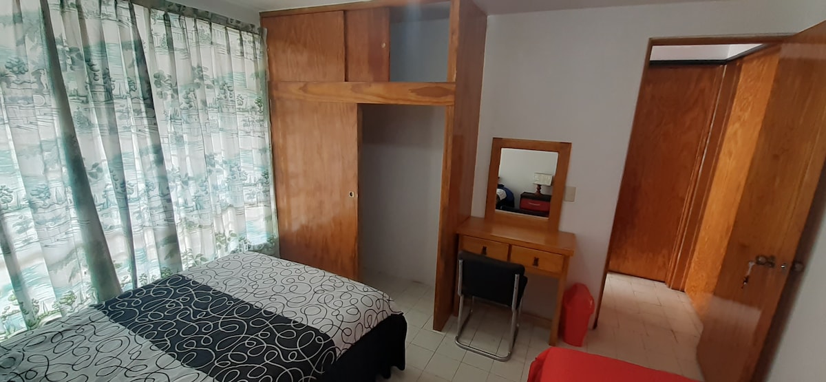 The room features two beds, one with a dark patterned cover and the other with a solid red cover. A wooden dresser is positioned against the wall, complemented by a small chair and a mirror. Natural light enters through a window dressed with light curtains.