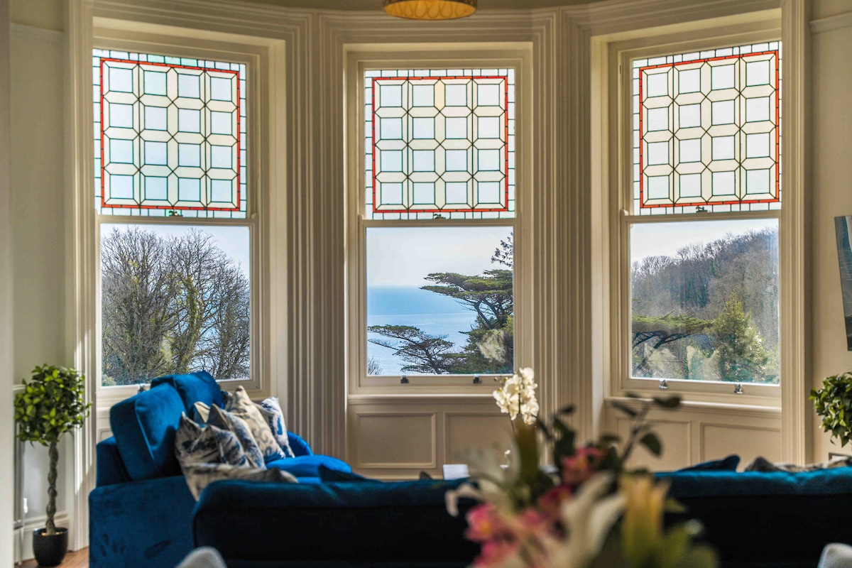 A bright seating area features a blue couch facing three large windows with intricate stained glass details. The windows offer views of the sea and surrounding trees, while a small potted plant adds a touch of greenery.
