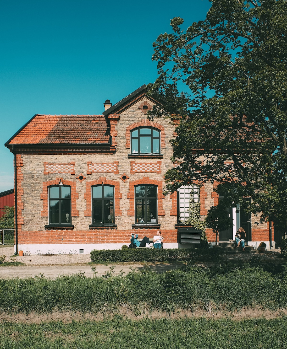 The exterior of a charming brick house is depicted, featuring tall windows with dark frames and decorative red detailing. A lush green lawn surrounds the property, and a large tree provides shade. Guests can be seen relaxing on the outdoor seating area.