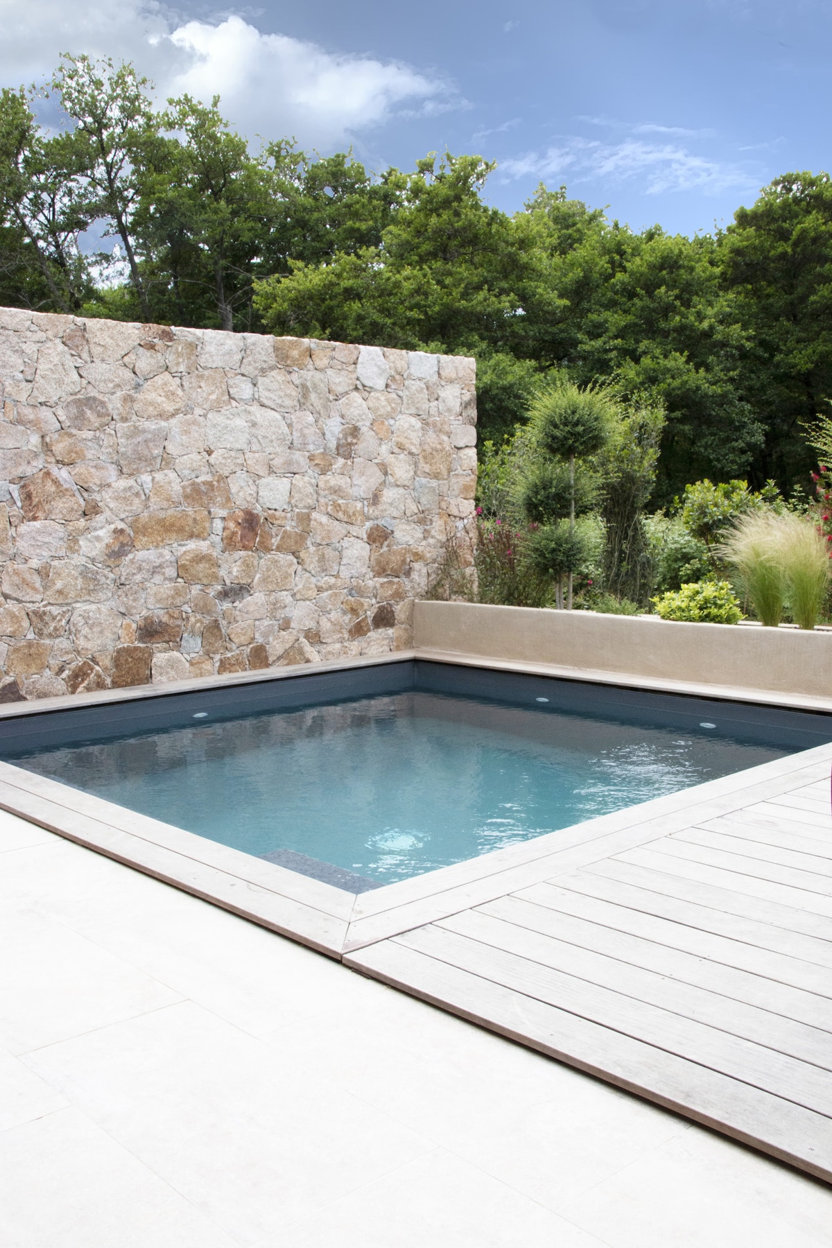 A private heated pool is set within a sleek wooden deck, surrounded by a natural stone wall. Lush greenery in the background enhances the serene environment, making it a peaceful space for relaxation.