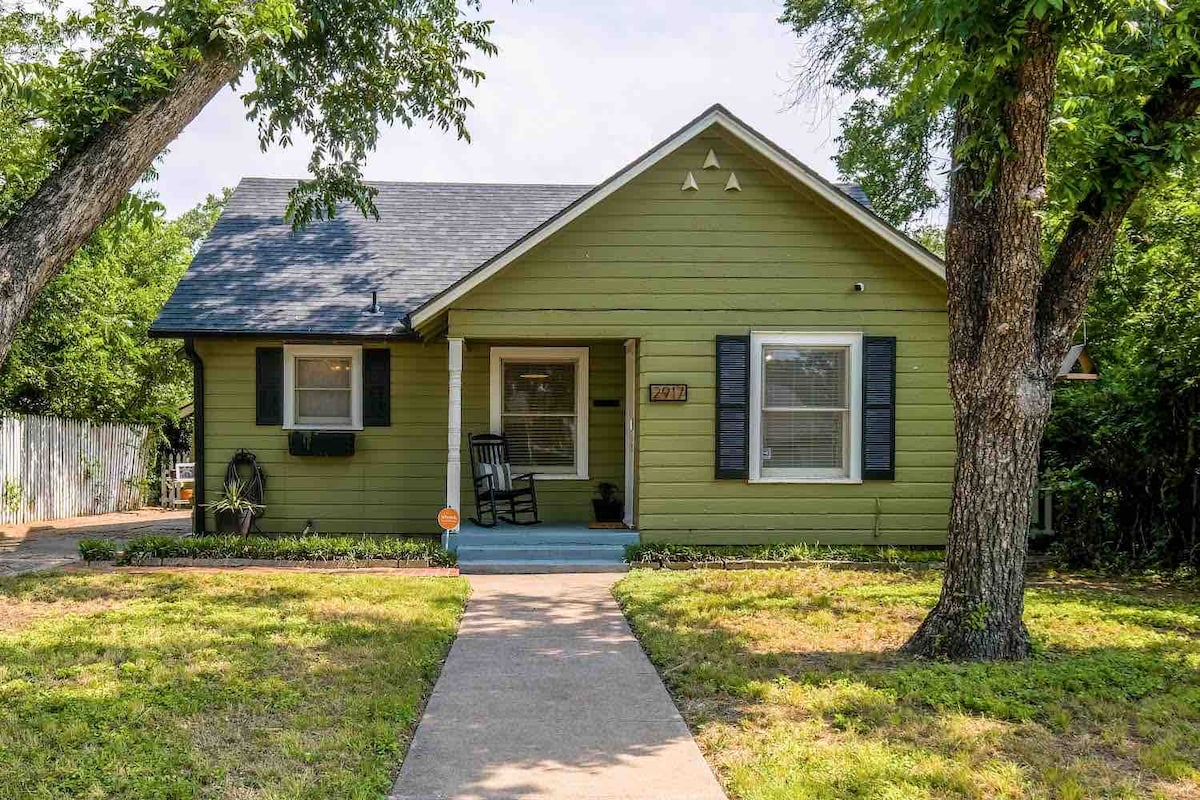 A charming green house is framed by two large trees, with a welcoming front porch featuring a rocking chair. A concrete pathway leads to the entrance, surrounded by a well-maintained lawn and landscaping. Air conditioning unit is visible on the left side of the house.