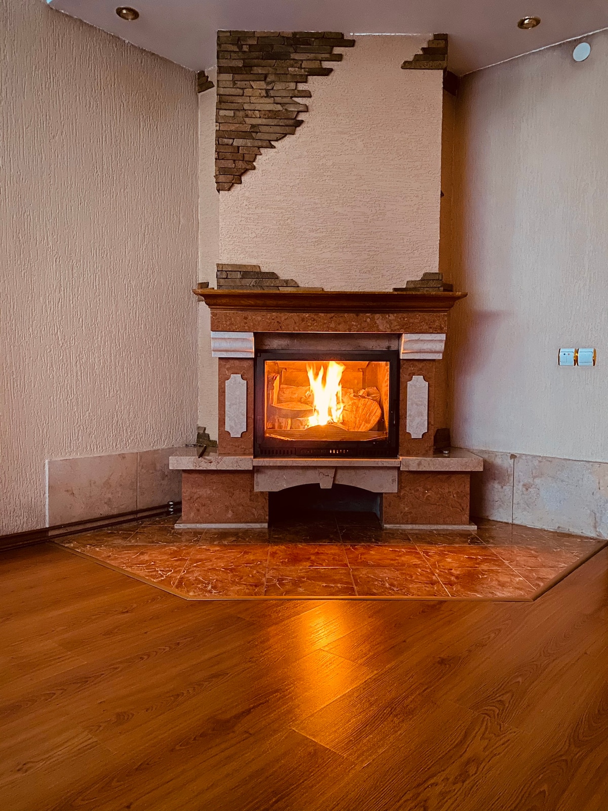 A warm fireplace is located in the corner of a room, featuring a stone and brick surround. Flames can be seen inside the stove, creating a cozy ambiance. The flooring is made of polished wood, complementing the inviting setting of the space.