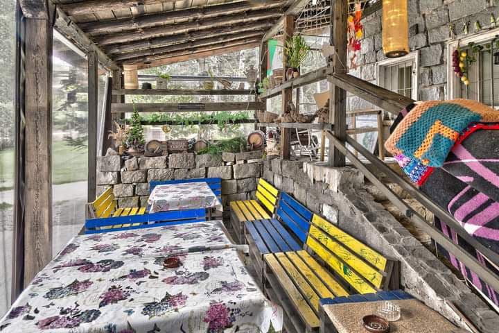 A covered patio area features wooden seating in yellow and blue, accompanied by tables adorned with floral tablecloths. The rustic stone wall provides a natural backdrop, while greenery is visible in the surroundings, contributing to a relaxed atmosphere.