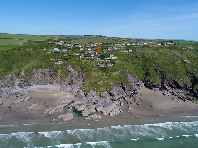 An aerial view showcases the coastal setting of Freathy, with the chalet highlighted in orange among lush greenery. The rugged cliffs lead to sandy beaches bordered by gentle waves, while the expansive sky reveals a hint of clouds, providing a serene backdrop to the picturesque landscape.