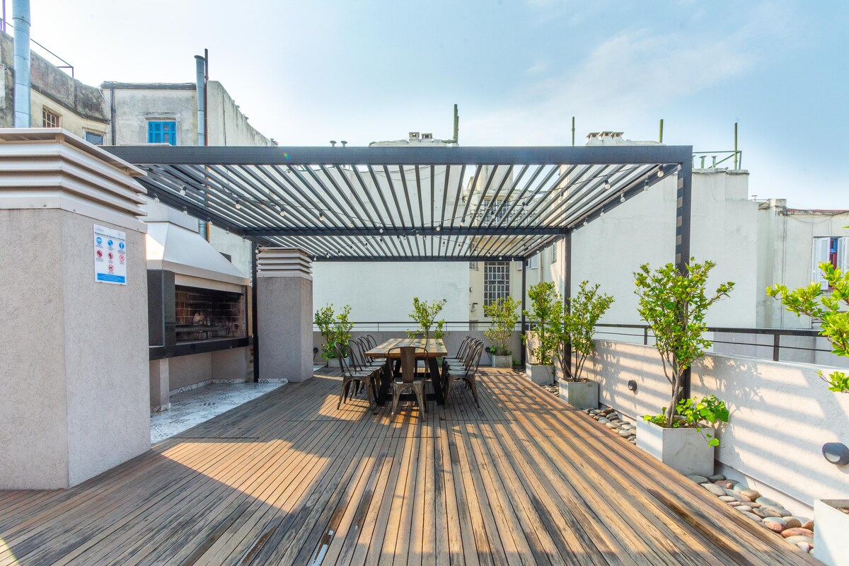 An open rooftop terrace features wooden decking and a dining table surrounded by several potted plants. A shaded area provides cover for a barbecue grill, creating a versatile space for gatherings and relaxation.
