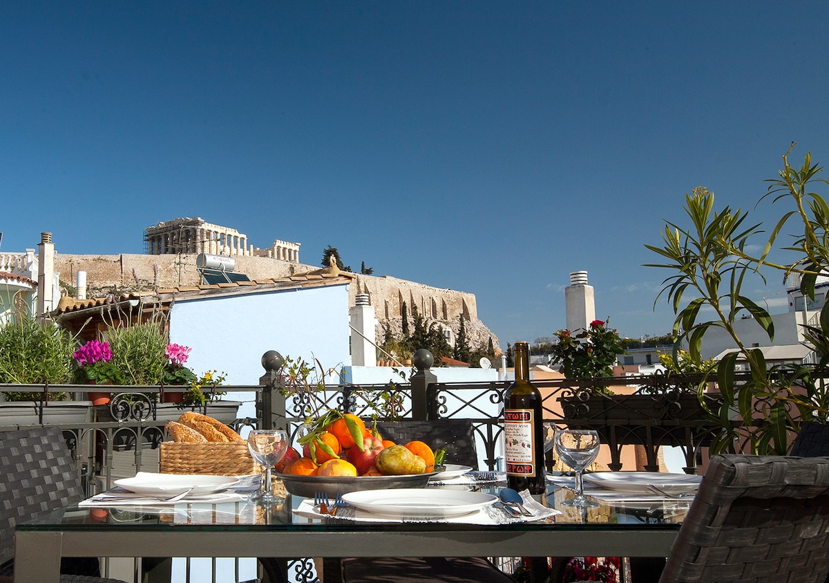 A dining table is set on a private roof garden terrace, featuring a variety of fresh fruits and a bottle of wine. In the background, the Acropolis and Parthenon are clearly visible against a bright blue sky, surrounded by greenery and colorful flowers.