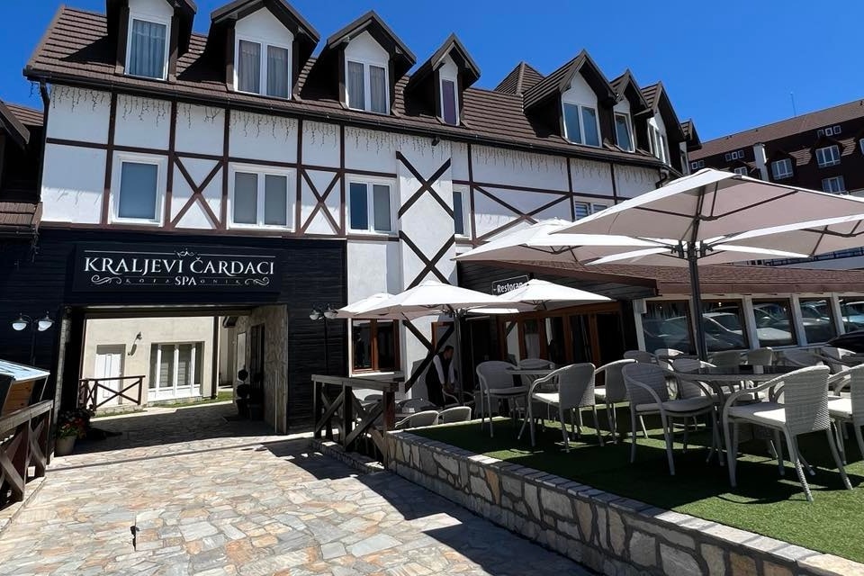 A modern building with a mix of dark wood and white exterior panels is featured. Outdoor seating areas with umbrella-covered tables and chairs are visible, accompanied by a paved pathway leading to the entrance. Clear blue skies provide a bright backdrop.
