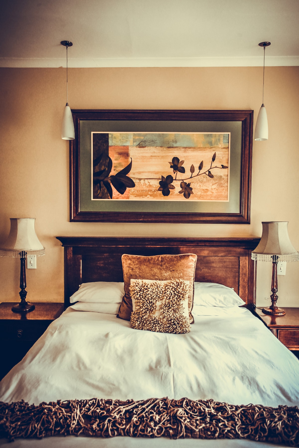 A comfortable sleeping area features a wooden bed with a neutral-colored blanket. Two bedside lamps are positioned beside the bed, and a decorative cushion adds an accent. A framed artwork above the headboard depicts floral motifs and warm tones, enhancing the overall aesthetic.