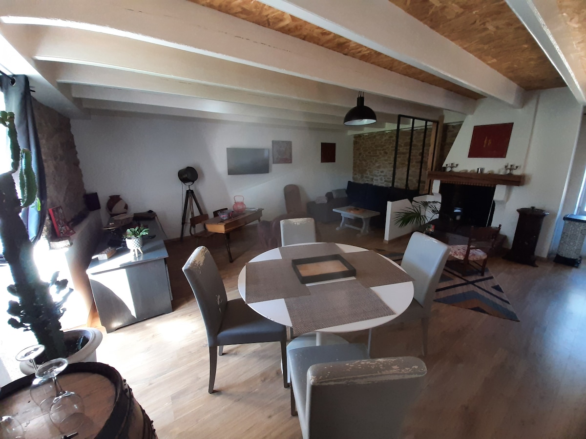 A spacious living area is shown with wooden flooring and exposed beams. A round dining table is surrounded by four chairs. A comfortable seating arrangement is positioned in the corner, with a decorative rug underneath. A fireplace and windows allow natural light into the space.