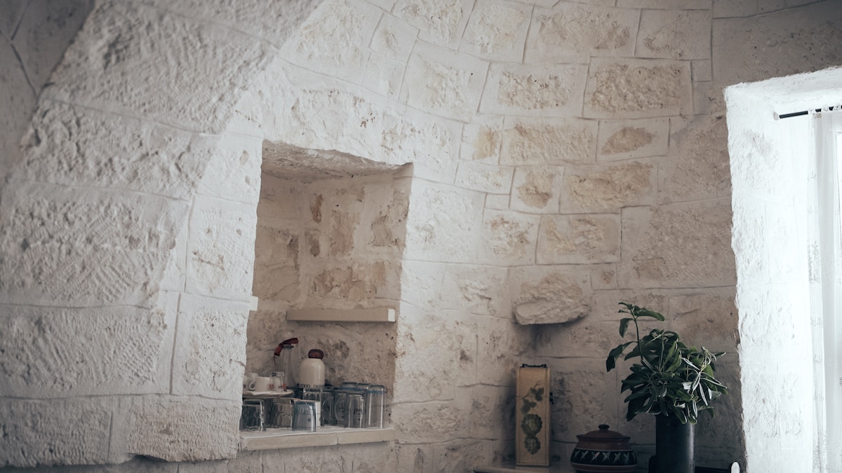The interior showcases natural stone walls with textured finishes, arched openings, and a shelf displaying glass jars and pottery. A small green plant adds a touch of nature, while a nearby window allows soft light to illuminate the space.