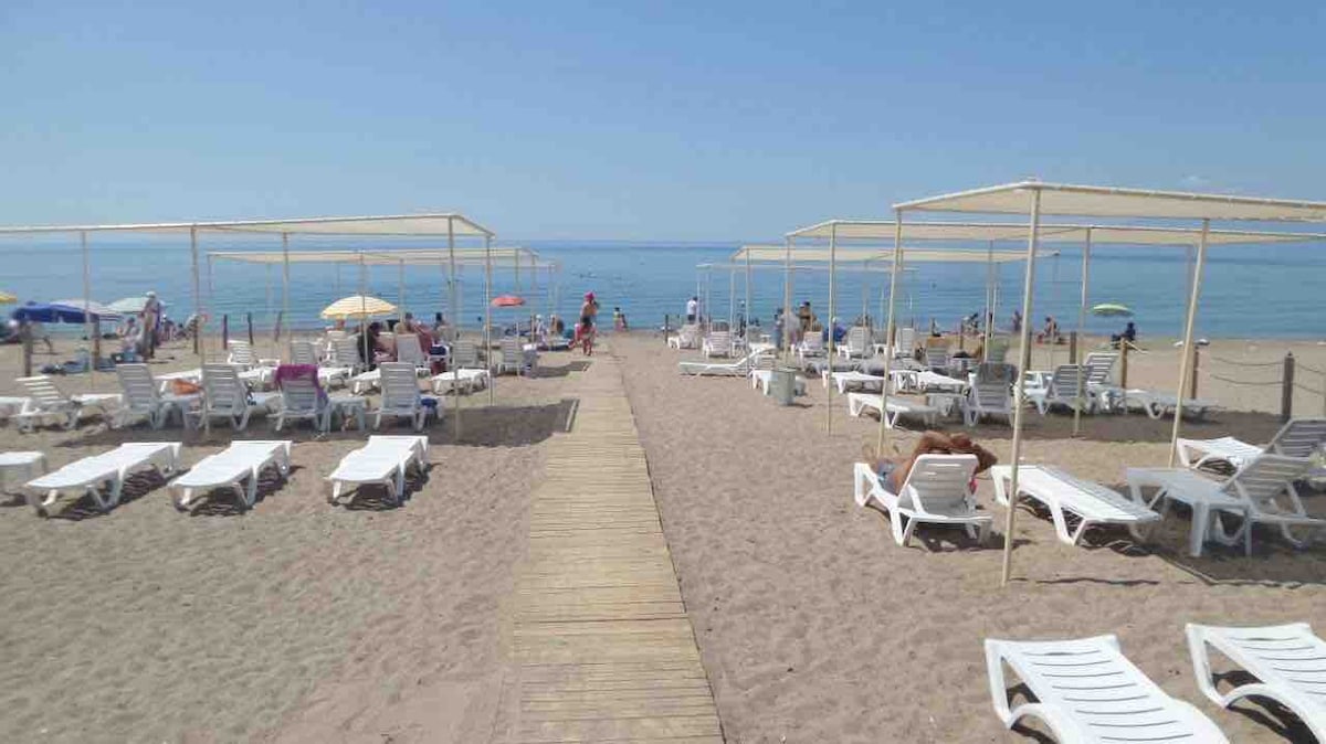 A sandy beach features numerous lounge chairs arranged under shaded canopies, leading to the calm sea. Sunbathers are seen relaxing in the distance, with colorful umbrellas scattered throughout the area. A wooden walkway guides visitors from the shore to the seating area.