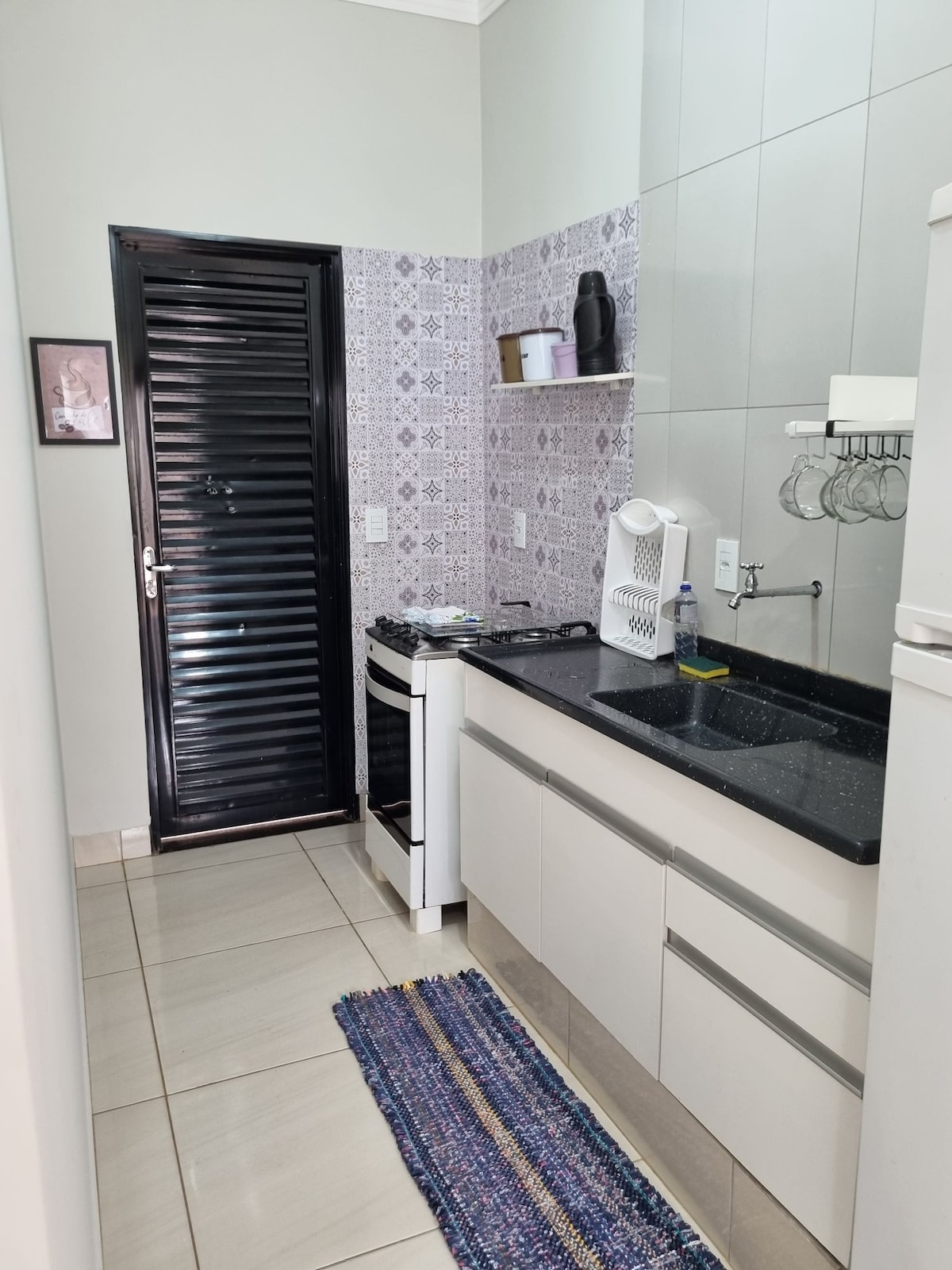 A modern kitchen is presented with sleek cabinetry and a dark countertop. Featured are a gas stove and a sink, with dishes and utensils arranged on the countertop. A decorative rug adds color to the tiled floor, and a closed black door is visible.