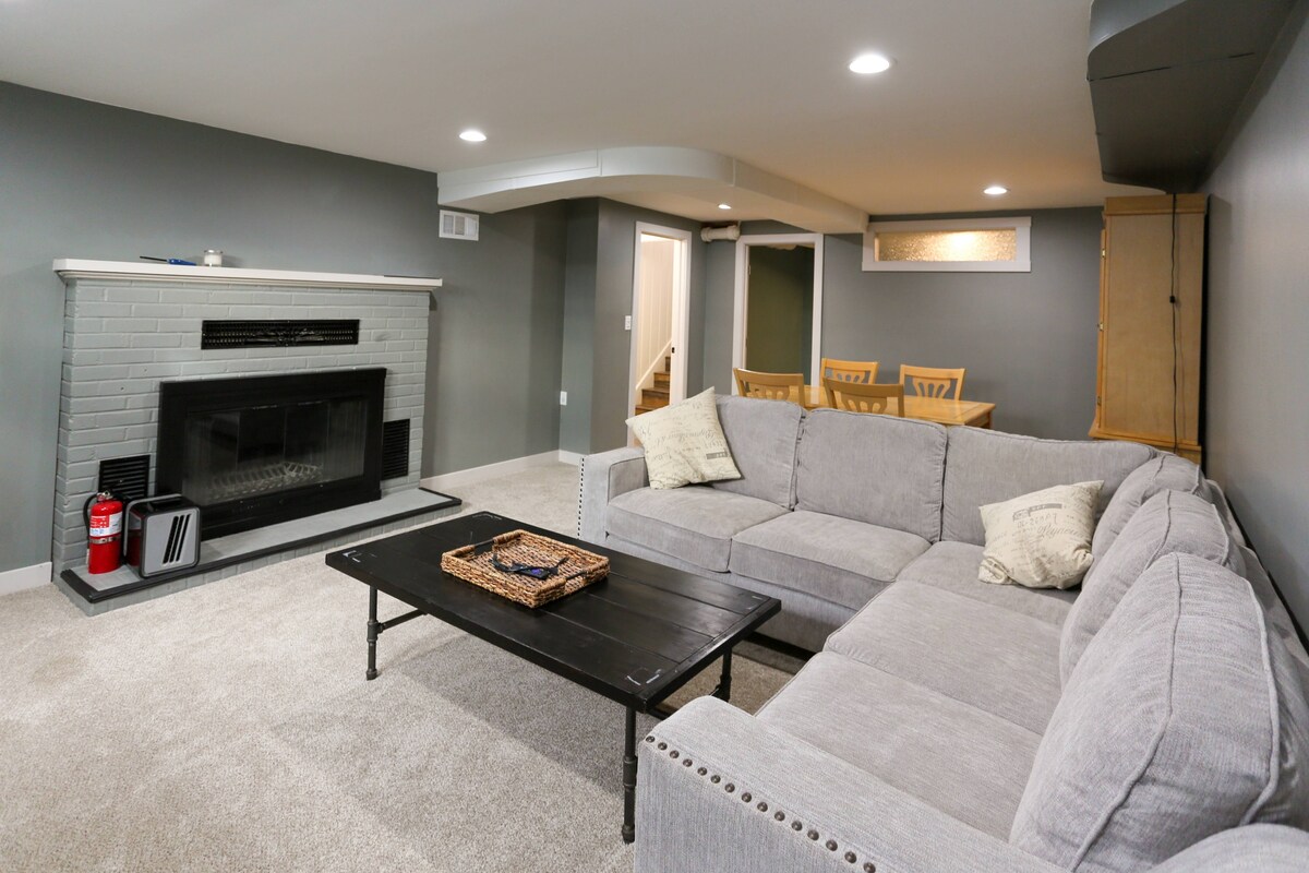 A spacious living area is shown, featuring a comfortable sectional sofa arranged around a dark coffee table. A fireplace is positioned on one side, complemented by light gray walls and a soft carpet. A dining area with wooden chairs is visible in the background.