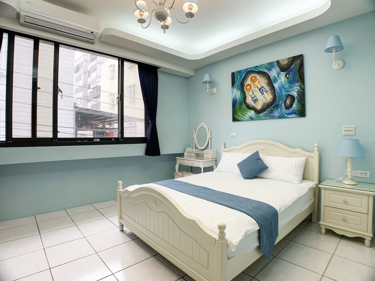 The room features a spacious bed dressed in white linens and a decorative blue throw. A large window allows natural light to fill the space, complemented by dark curtains. An artistic wall painting adds color, while bedside tables and a vanity enhance functionality.