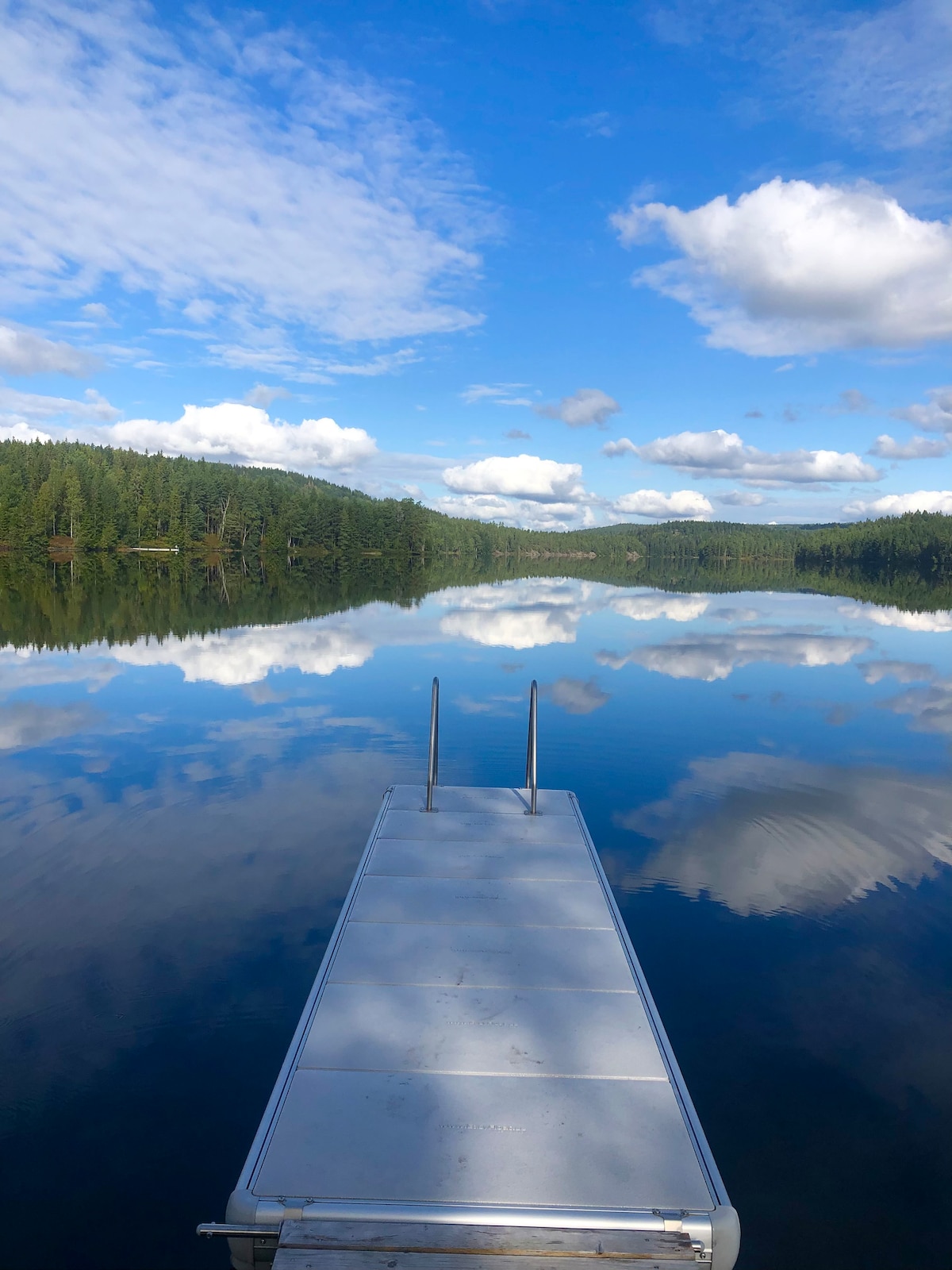 A jetty extends into calm waters, reflecting the blue sky and scattered clouds above. Lush greenery surrounds the lake, creating a serene backdrop. The stillness of the water showcases the peaceful natural environment.