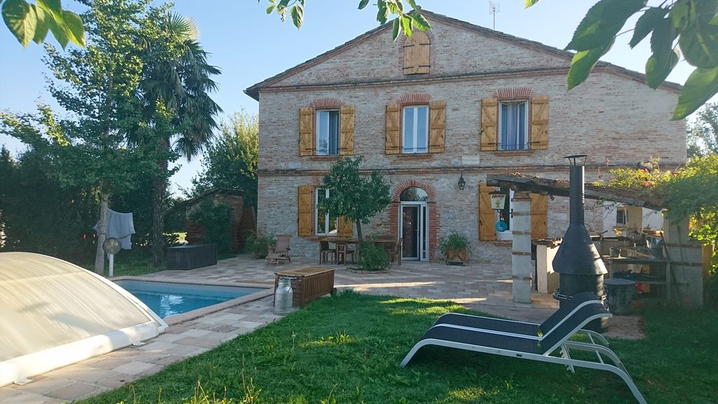 A brick farmhouse is surrounded by greenery, featuring yellow shutters and a covered pool. Outdoor seating and a barbecue area can be seen alongside a lounge chair on the lawn. Sunlight casts a warm glow over the peaceful garden setting.