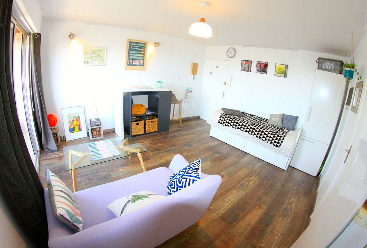 A spacious studio is showcased with wooden flooring and bright walls. A comfortable purple sofa and a black-and-white patterned bed are positioned in the living area. Natural light fills the room through a large window, complemented by decorative artwork and a coffee table.