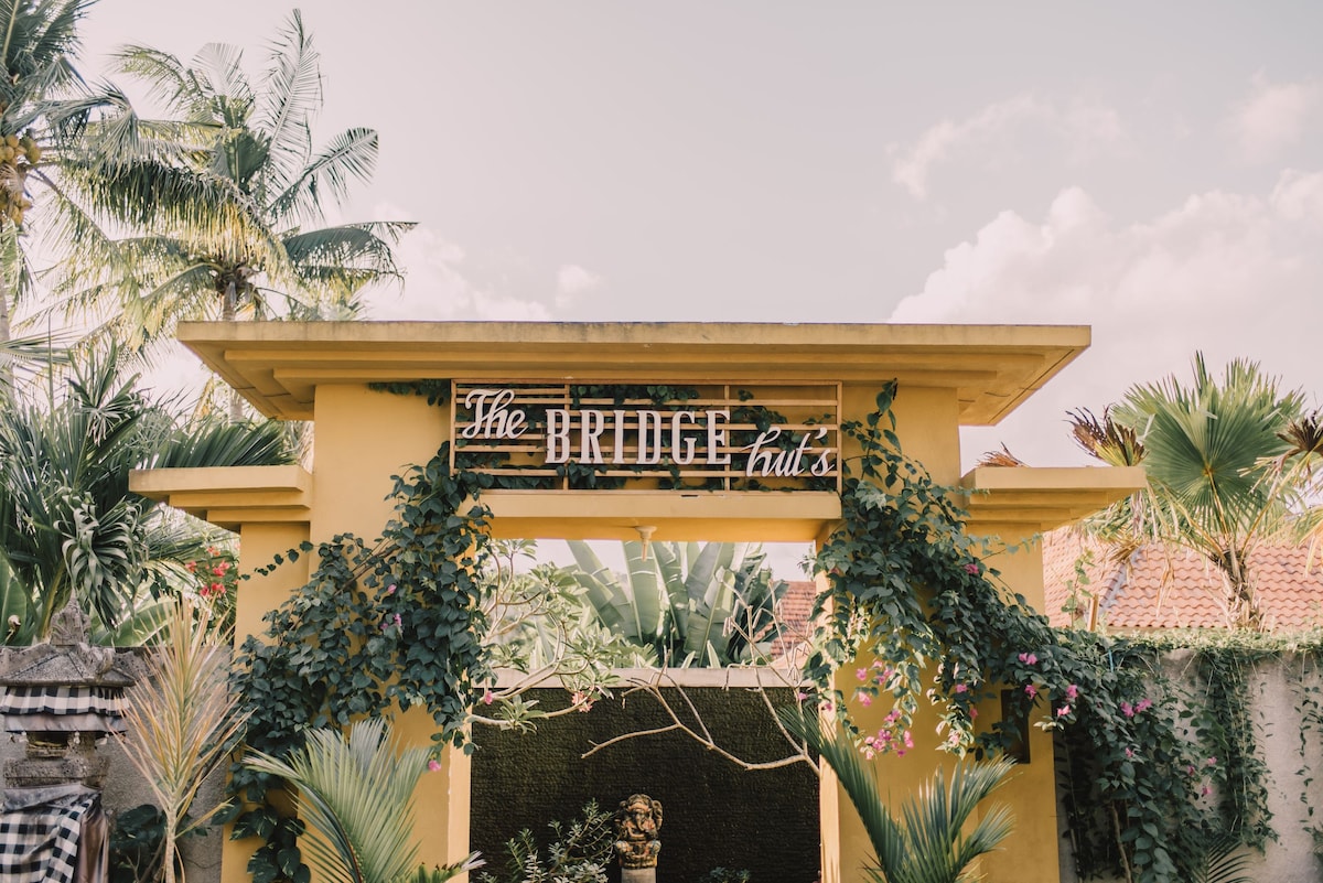 A welcoming entrance to The Bridge Huts is framed by lush greenery and vibrant flowering plants. The sign displaying 'The Bridge Huts' is prominently featured above an arched doorway, inviting guests into a tropical atmosphere.