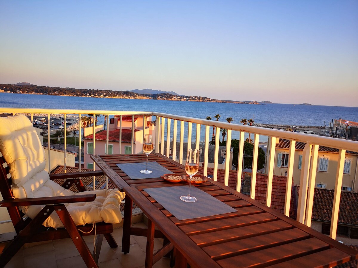 A spacious balcony is featured, showcasing a wooden dining table set for two with glasses and a small dish. Scenic views of the bay and distant coastline are visible during sunset, enhancing the inviting outdoor space.