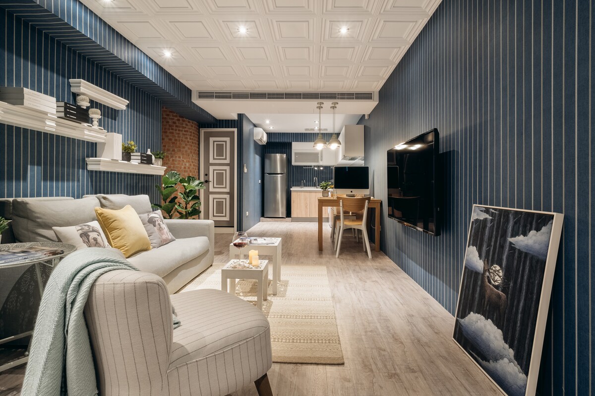A modern living space is highlighted with blue-striped walls and stylish decor. A comfortable sofa faces a mounted television, while a small dining area is visible in the background. Green plants add a touch of nature, complemented by warm lighting from ceiling fixtures.