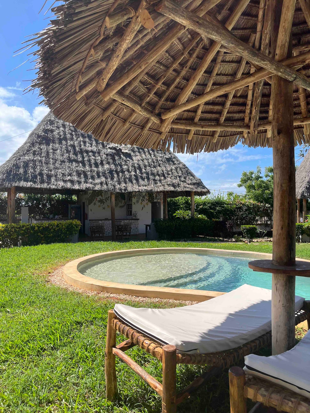 A serene outdoor area features a circular swimming pool surrounded by lush greenery. A thatched-roof gazebo provides shade over a comfortable lounge chair. The bungalows are visible in the background, blending harmoniously with the garden landscape.