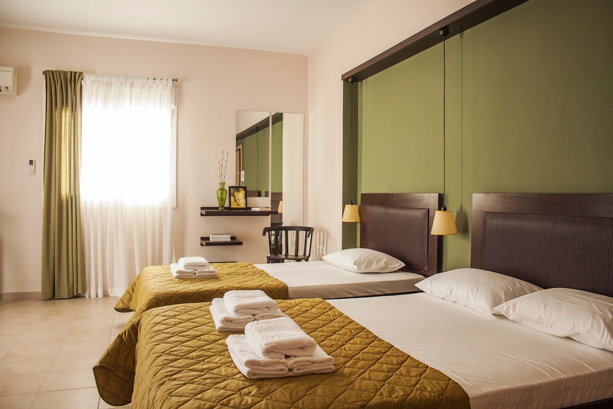 An inviting guest room features two spacious beds with neatly arranged linens. Soft natural light enters through a window, complemented by sheer curtains. A minimalist mirror and a small shelf are mounted on a neutral wall, while a cozy chair adds to the room's functionality.