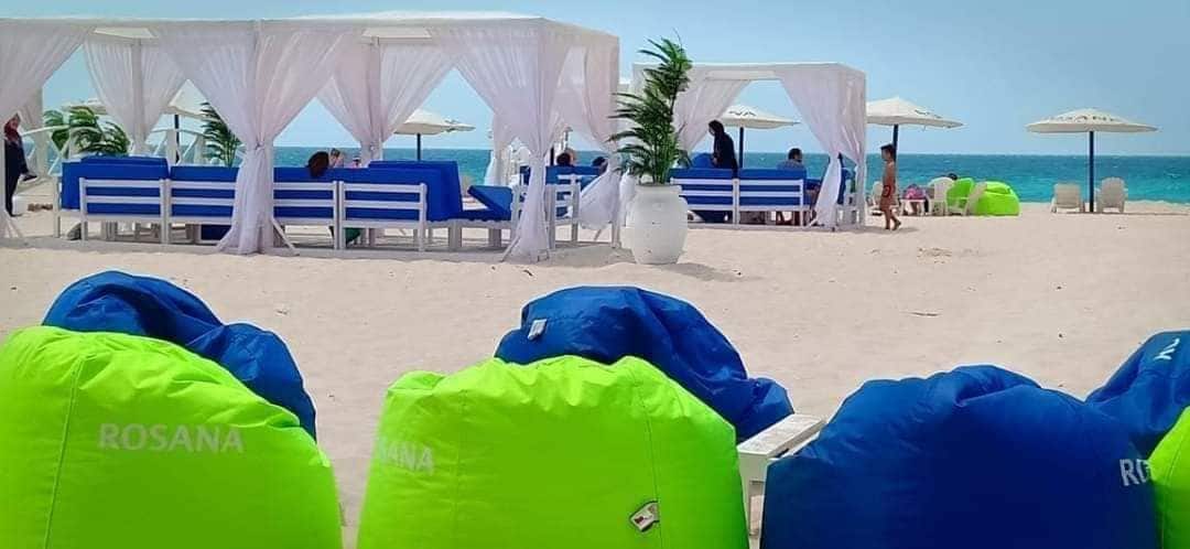 Colorful bean bags in shades of blue and green are arranged on a sandy beach. White canopies and lounge seating provide shaded areas nearby. Umbrellas are visible in the background, overlooking the ocean, creating a relaxed seaside atmosphere.