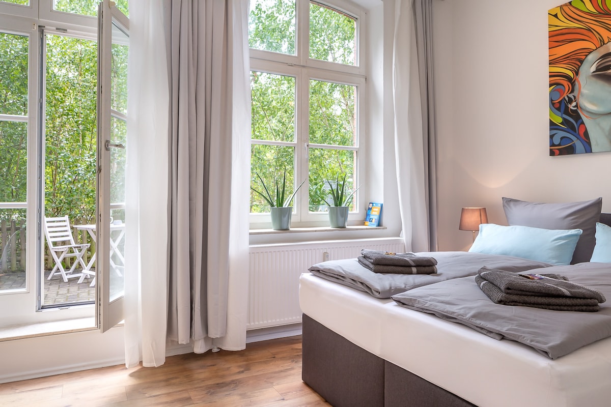 A well-lit bedroom showcases a spacious bed with smooth linens and neatly arranged towels. Large windows invite natural light and offer a view of a serene terrace. Green potted plants add a touch of nature, enhancing the calming ambiance of the room.