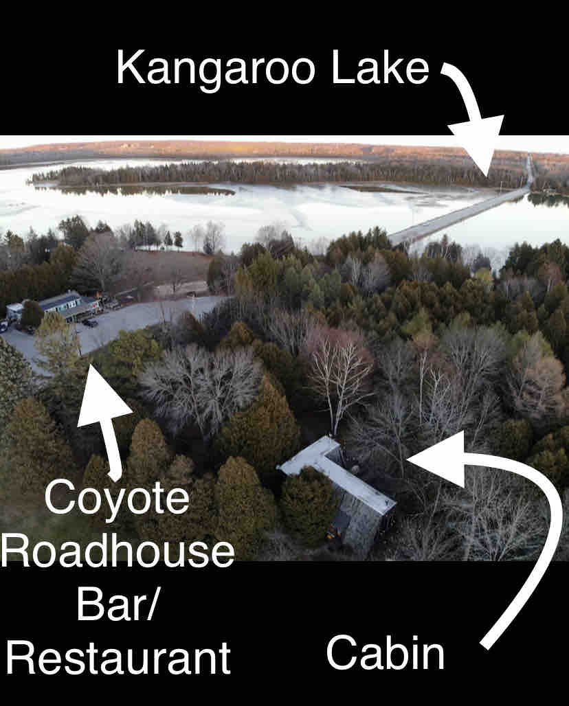 An aerial view captures the cabin nestled among trees, with Kangaroo Lake visible nearby. A labeled path indicates the location of Coyote Roadhouse Bar/Restaurant. The serene landscape showcases the surrounding nature and proximity to water, highlighting the secluded setting.