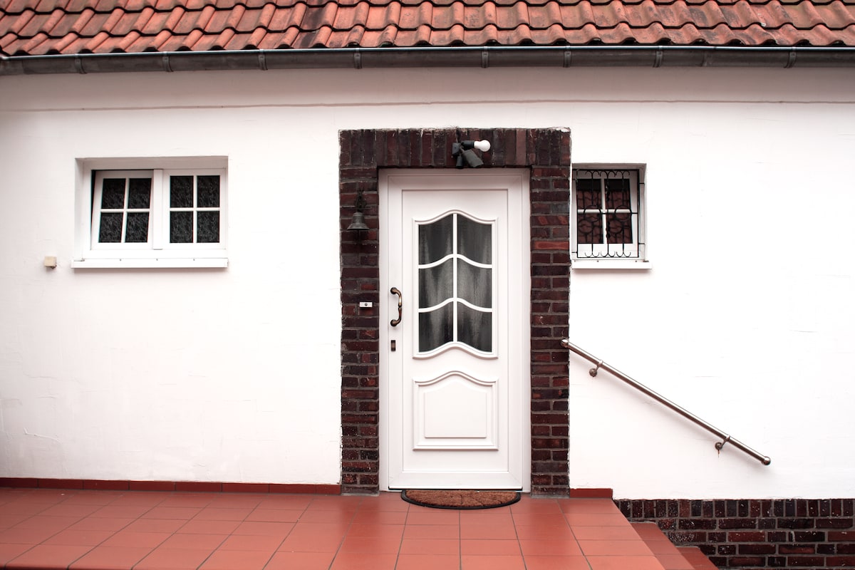 The entrance of the property is framed by a brick wall and features a white door with decorative glass panels. A small mat is placed in front, and a handrail provides support for accessing the steps. Two windows are located on either side of the door.