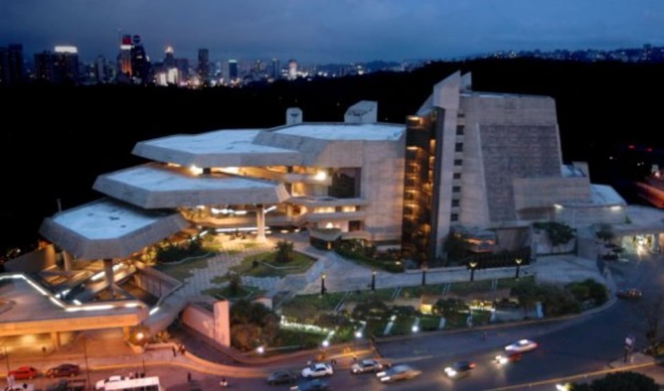 A modern architectural structure is depicted, showcasing unique geometric shapes and concrete finishes. Surrounding greenery is visible, with pathways leading up to the entrance. The city skyline is faintly illuminated in the background, hinting at the vibrant atmosphere of downtown Caracas during twilight.