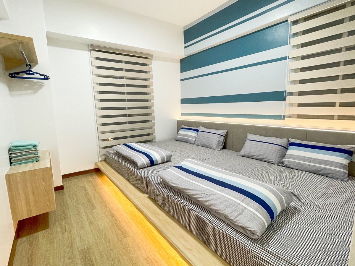 A well-lit bedroom features two king size beds with simple bedding, positioned against a striped accent wall. Soft ambient lighting highlights the wooden floor, while shelves are present for convenience. Window coverings allow for light control, maintaining brightness and privacy.