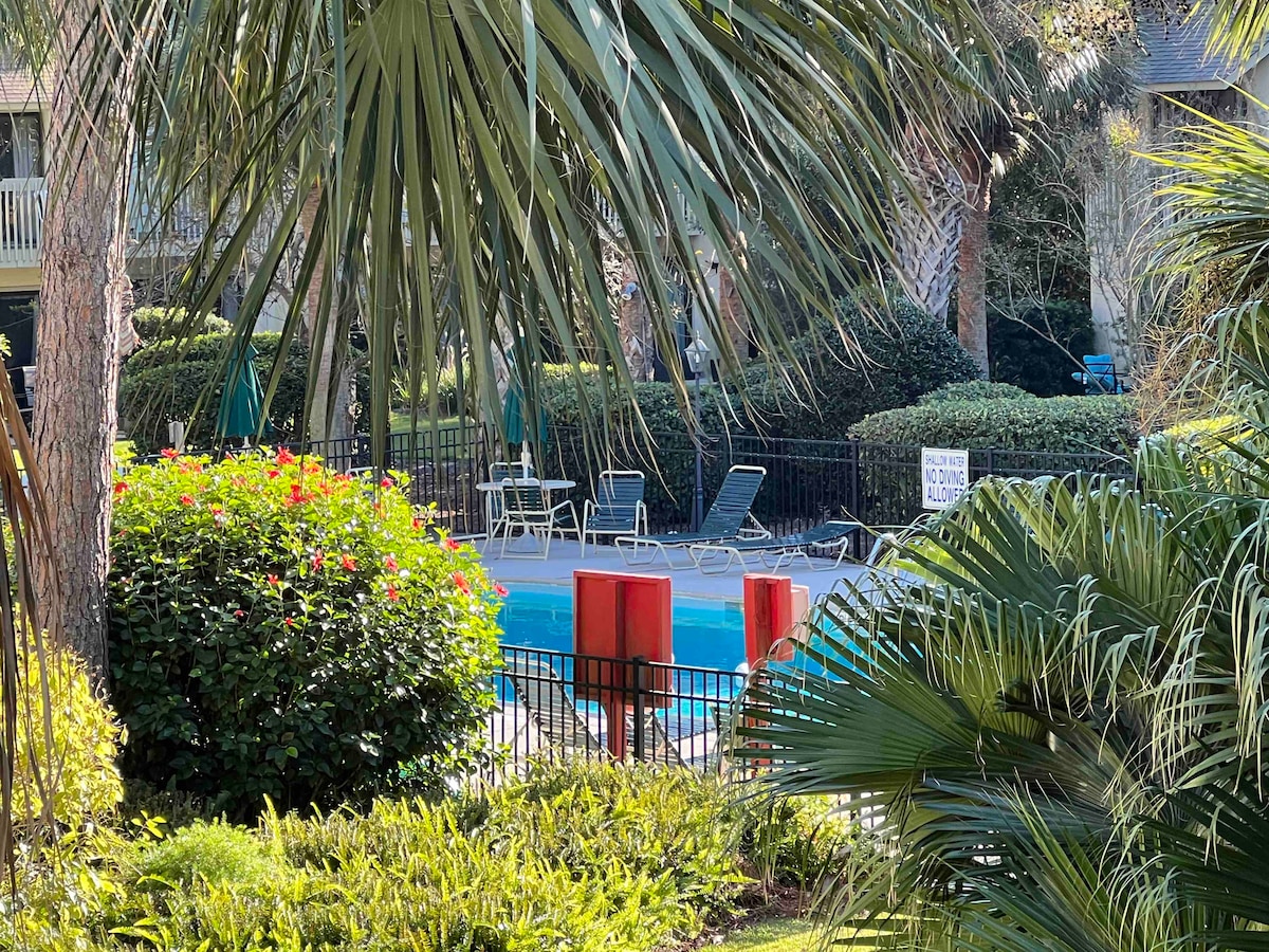 A glimpse of the pool area is framed by lush palm trees and vibrant greenery. Lounge chairs are positioned around the pool, with a protective fence visible in the background. Colorful flowers add to the serene setting, enhancing the overall outdoor ambiance.