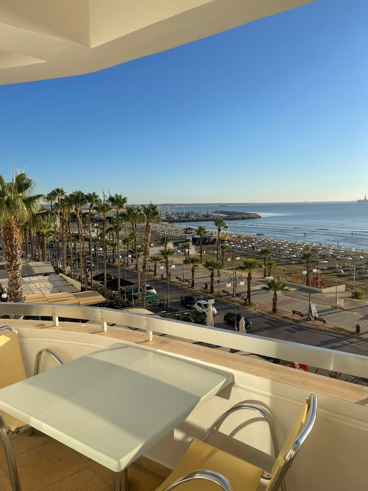 A balcony view features a clear blue sky overlooking a sandy beach lined with palm trees. Sun loungers are placed along the shore, and boats can be seen in the water, creating a serene coastal atmosphere.