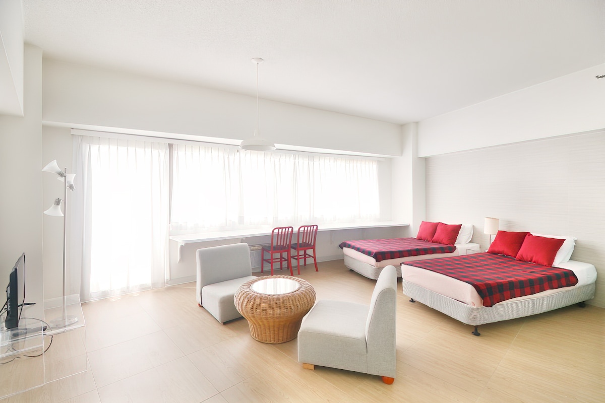A bright and airy room features two spacious beds, each adorned with red and white accents. Natural light flows in from large windows dressed with sheer curtains. A round, woven coffee table is complemented by modern seating, alongside a TV placed on a glass stand.