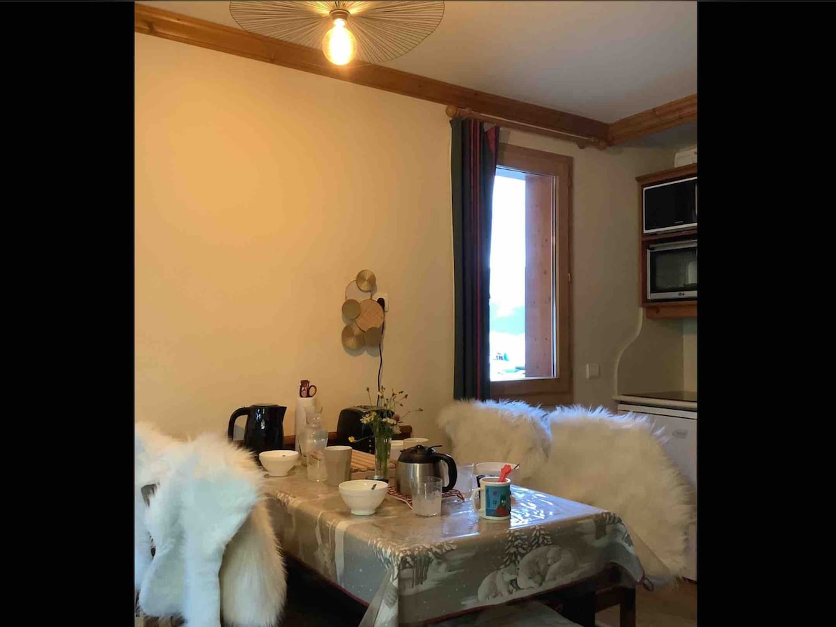 A dining area is featured with a table set for meals, accompanied by several chairs with soft faux fur covers. A window provides natural light, with minimal decor on the wall nearby. Kitchen appliances are visible in the background.