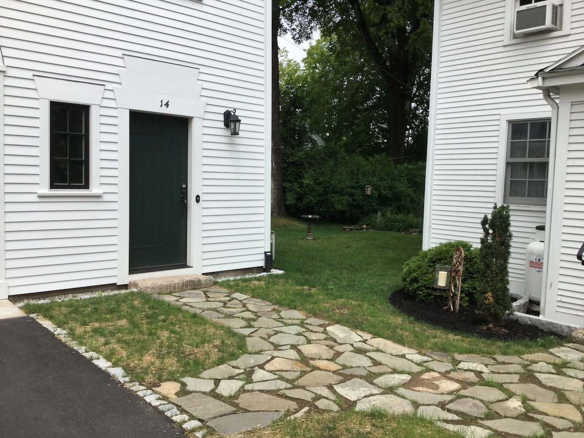 A stone pathway leads to a green door of a two-story white building. Lush greenery surrounds the entrance, with a well-maintained lawn and shrubs visible. Nearby, an adjacent building is seen, enhancing the serene outdoor setting.