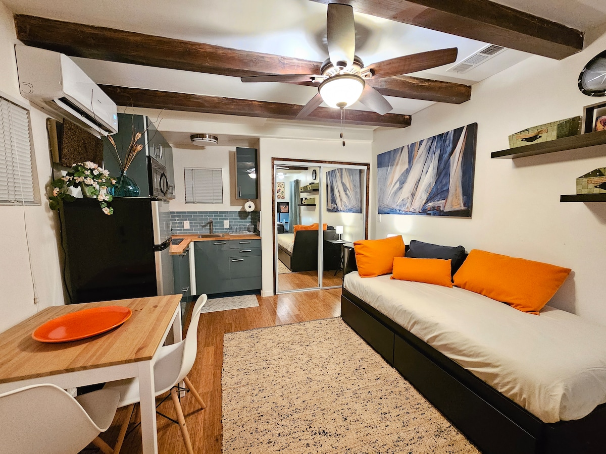 This stylish studio features wooden beams overhead and a cozy seating area with bright orange and blue accents. A small dining table is present, while a spacious bed and mirrored wardrobe reflect light, creating a welcoming atmosphere. The kitchen area is partially visible in the background.