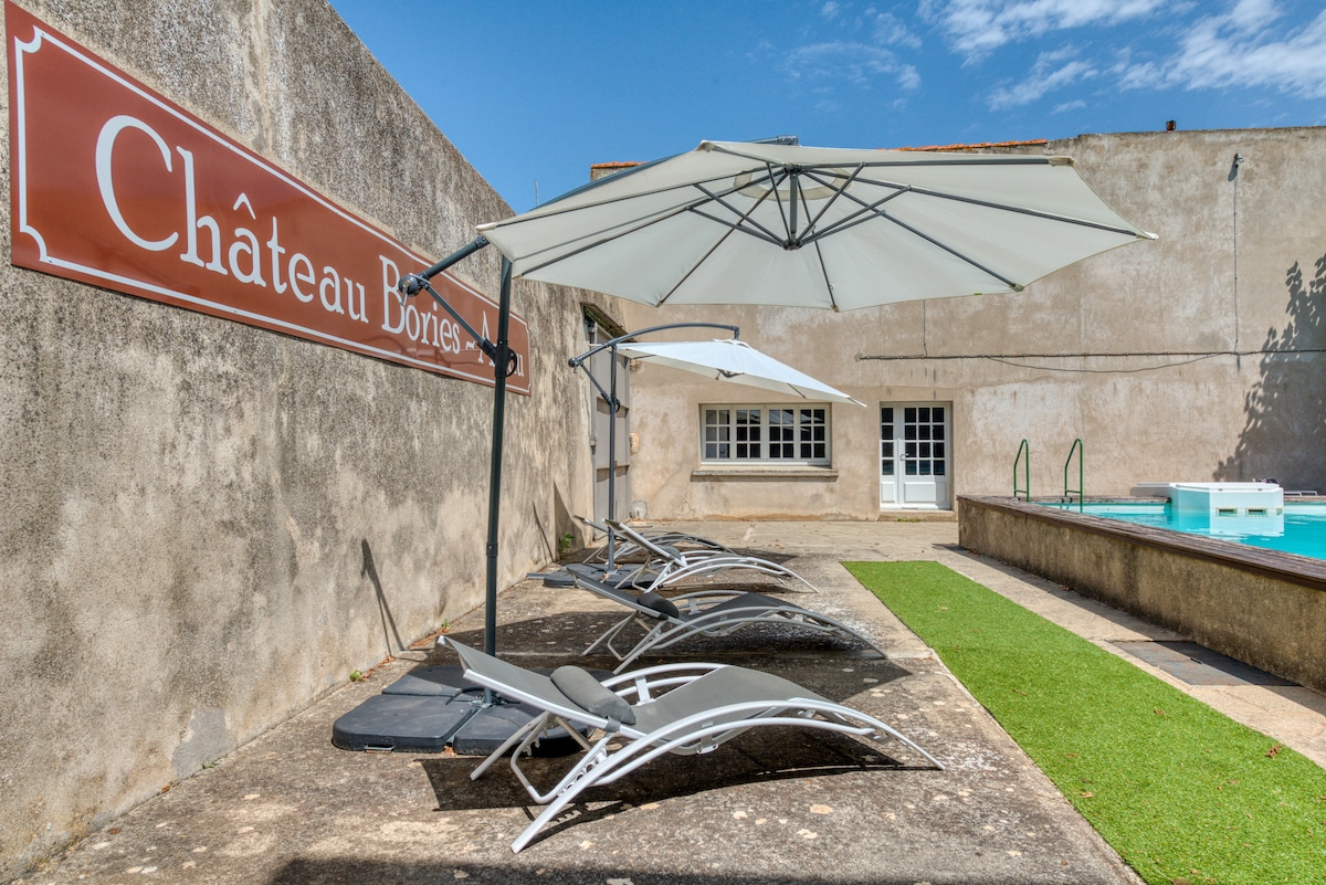 A sunlit outdoor area is showcased with four loungers arranged beneath a large umbrella. A sign for 'Château Bonies' is prominently displayed, while a small patch of green grass leads to the edge of a relaxing pool. The surroundings feature walls in muted tones.