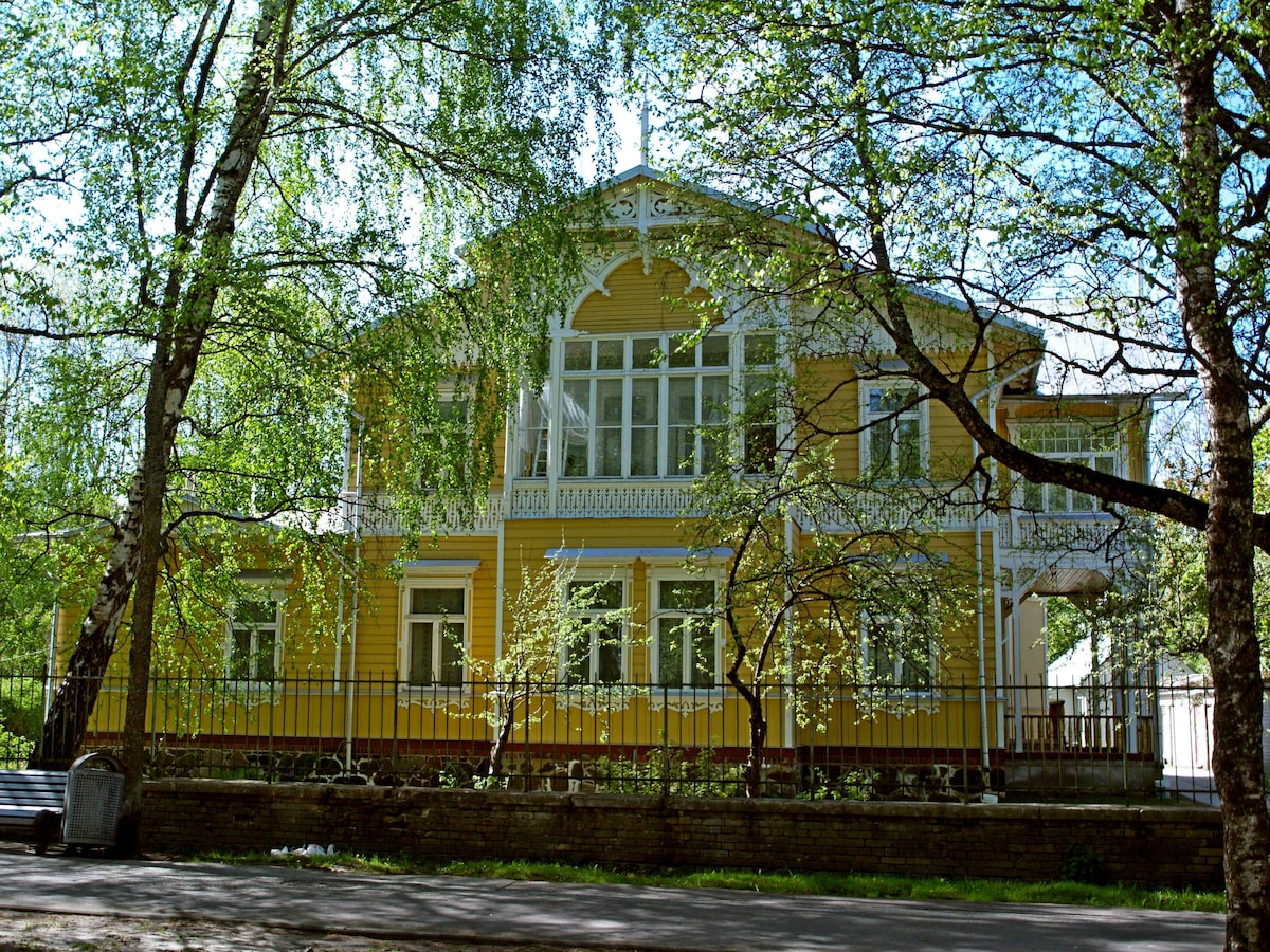 A historic yellow villa is surrounded by lush greenery, featuring large windows that reflect natural light. The decorative woodwork enhances the architectural details, and a charming balcony is visible above the entrance. The property is set back from the street, providing a sense of privacy.