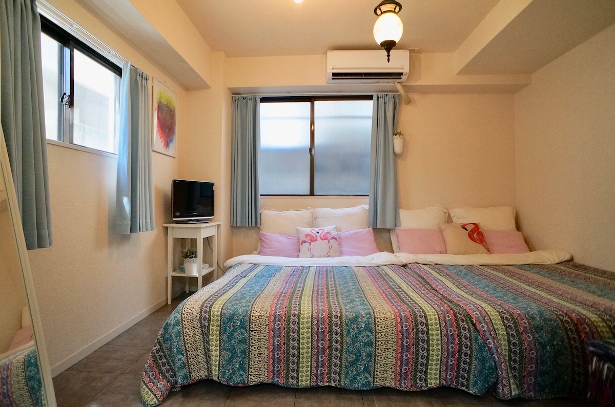 A spacious sleeping area features a large double bed covered with a colorful patterned blanket. Soft pillows are arranged atop the bed. Natural light enters through a large window framed with light blue curtains. A small TV and table are positioned nearby.