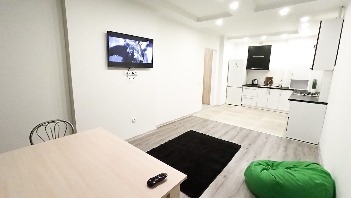 A modern studio apartment displays an open layout. A light wood dining table is positioned against the wall, accompanied by a comfortable chair. A green beanbag is located on the grey floor. A kitchenette featuring white cabinetry and essential appliances is visible in the background.